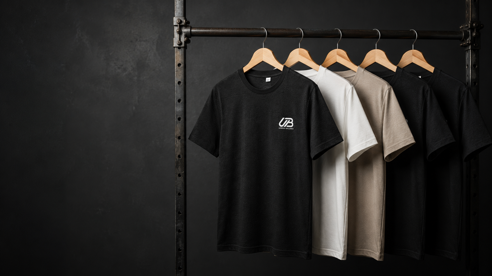 Five short-sleeve crewneck t-shirts in black, white, beige, and dark gray hanging on wooden hangers from a black metal rack against a dark textured wall.