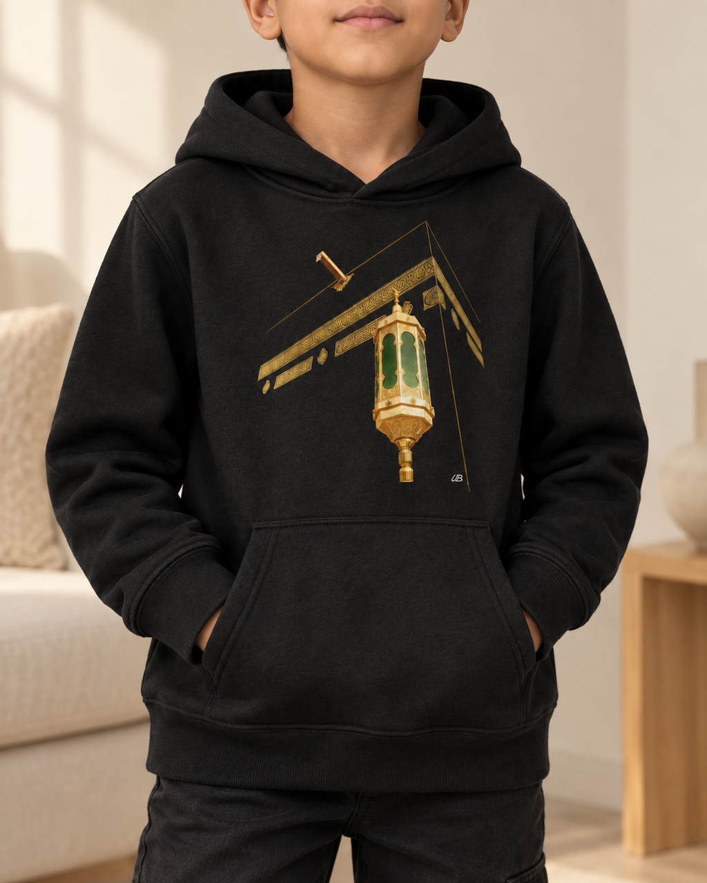 A child wearing a black pullover hoodie with a graphic print of a lantern and the Ka'bah, set against a softly blurred indoor background.