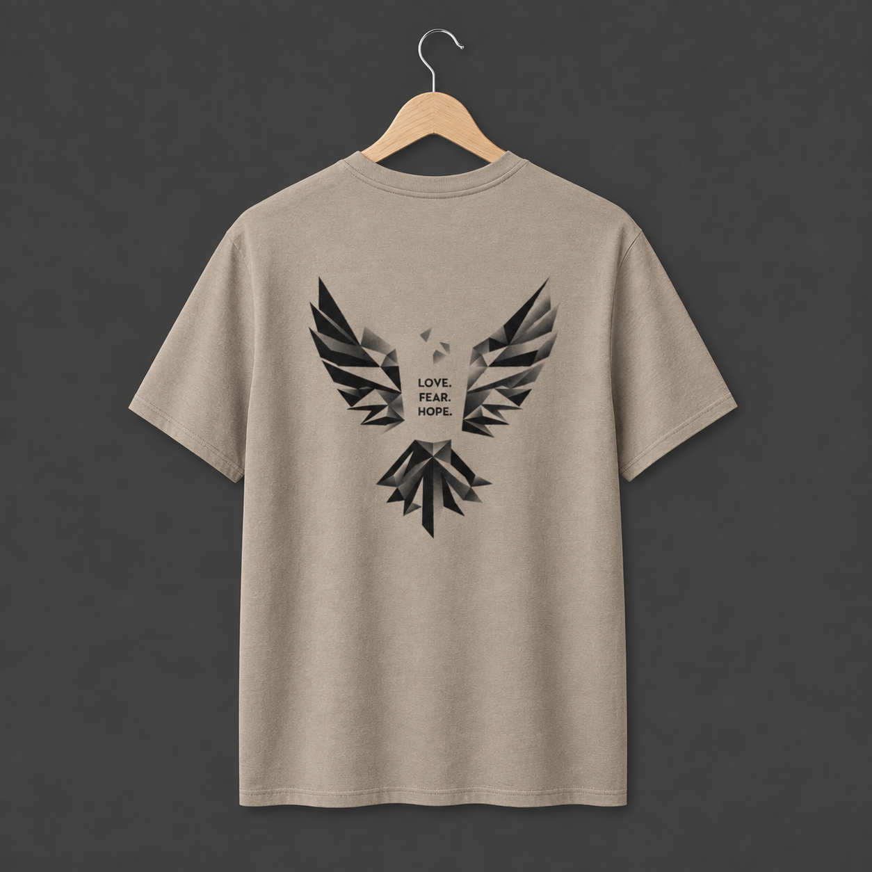 A dessert dust coloured short-sleeve t-shirt hanging on a wooden hanger. The back of the shirt features a large black abstract geometric bird graphic with the words 'LOVE. FEAR. HOPE.' in black in the middle. 