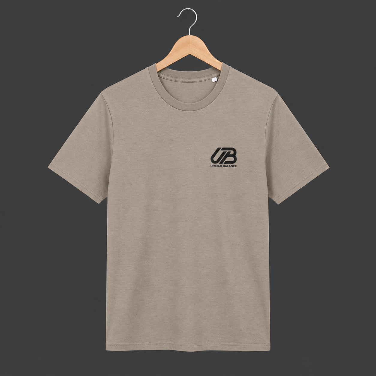 A dessert dust coloured short-sleeve crewneck t-shirt hanging on a wooden hanger. The shirt features a small black 'UB' logo on the left chest with 'Ummah Balance' printed underneath.