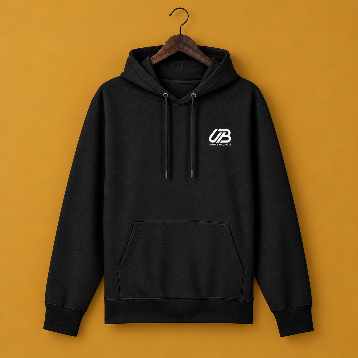 A black pullover hoodie with a white stylised 'UB UMMAH BALANCE' logo on the left chest, hanging on a wooden hanger against a solid mustard yellow background.