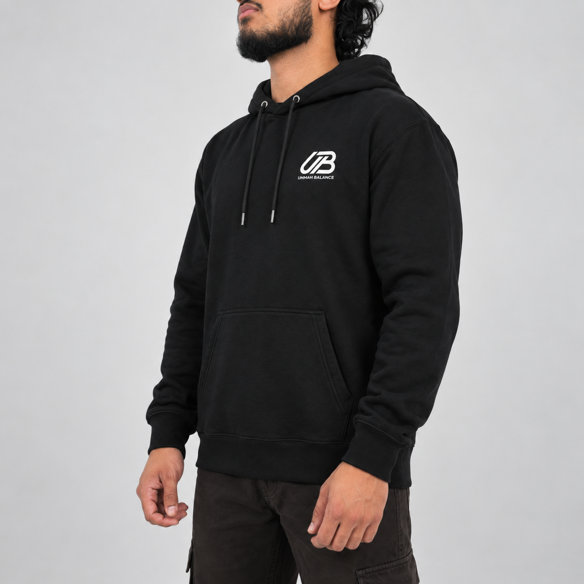 A man wearing a black pullover hoodie with a white 'UB UMMAH BALANCE' logo on the left chest, paired with dark cargo pants, photographed against a neutral grey studio background.