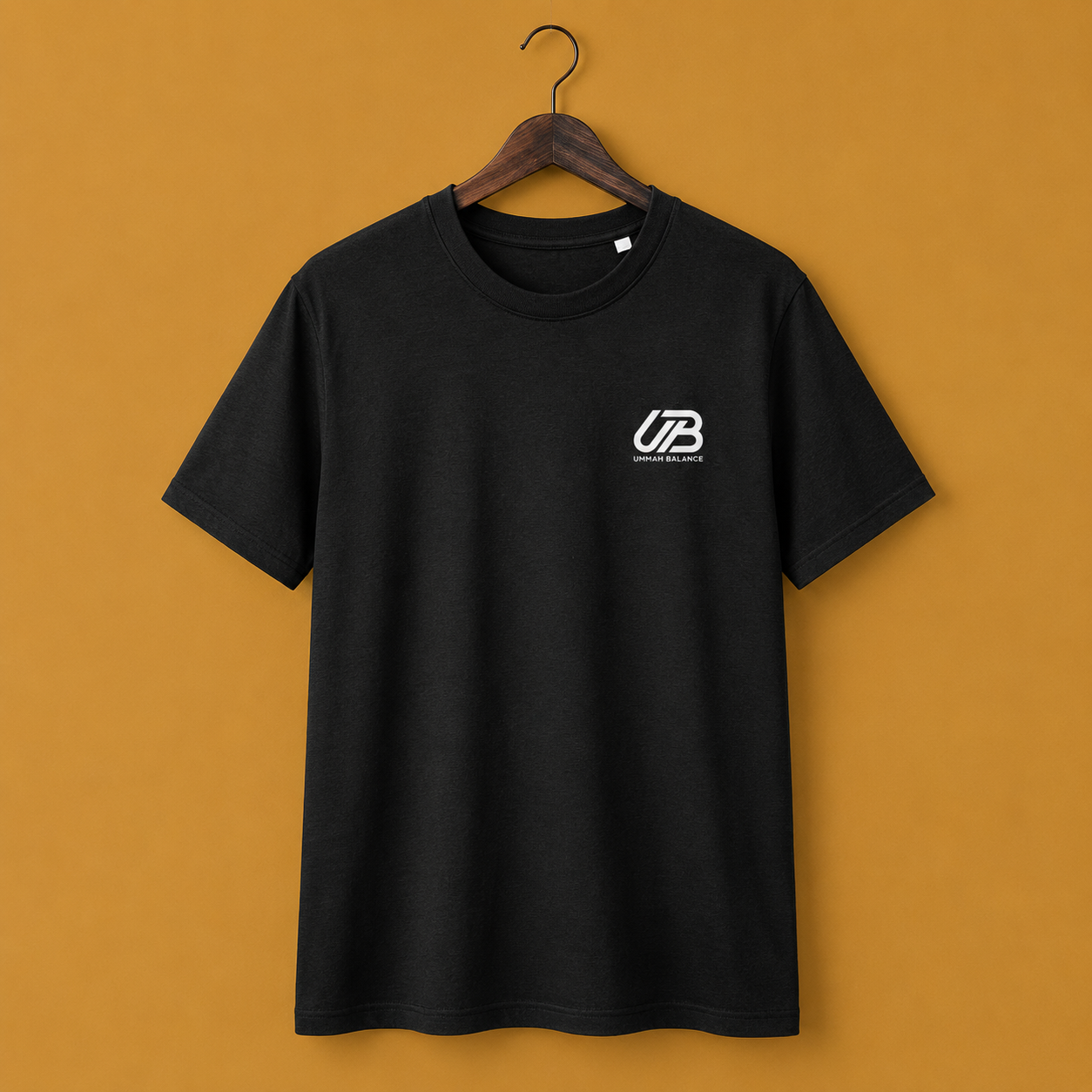 A black short-sleeve crewneck t-shirt hanging on a wooden hanger. The shirt features a small white 'UB' logo on the left chest with 'Ummah Balance' printed underneath.