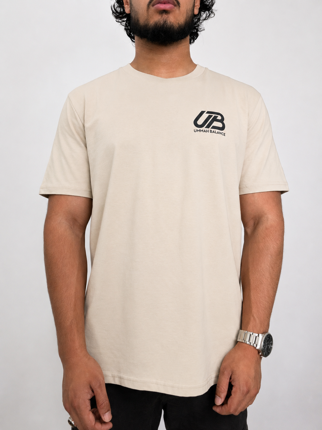 A man wearing a desert dust crew neck t-shirt with a black 'UB' logo and 'Ummah Balance' text on the left chest, paired with black pants, against a plain light grey background.