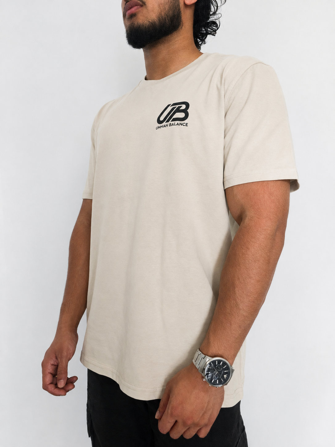 A man wearing a desrt dust short-sleeve t-shirt with a black 'UB' and 'Ummah Balance' logo on the left chest, paired with black pants and a silver watch, against a plain white background.