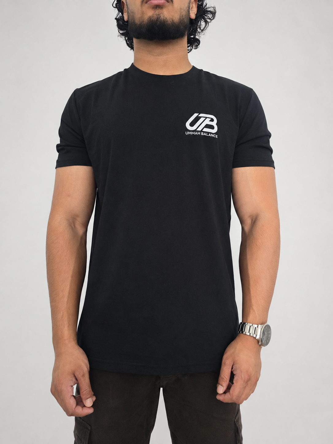A man wearing a black short-sleeve t-shirt with a white 'UB' and 'Ummah Balance" logo on the left chest, paired with dark brown pants, photographed against a neutral light grey background.