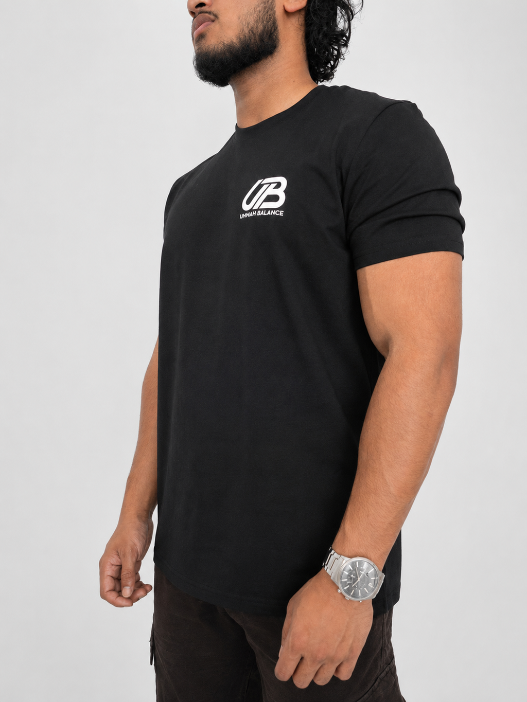A man wearing a black short-sleeve t-shirt with a white 'UB' logo and 'Ummah Balance' text on the left chest, shown from the waist up against a light grey background.
