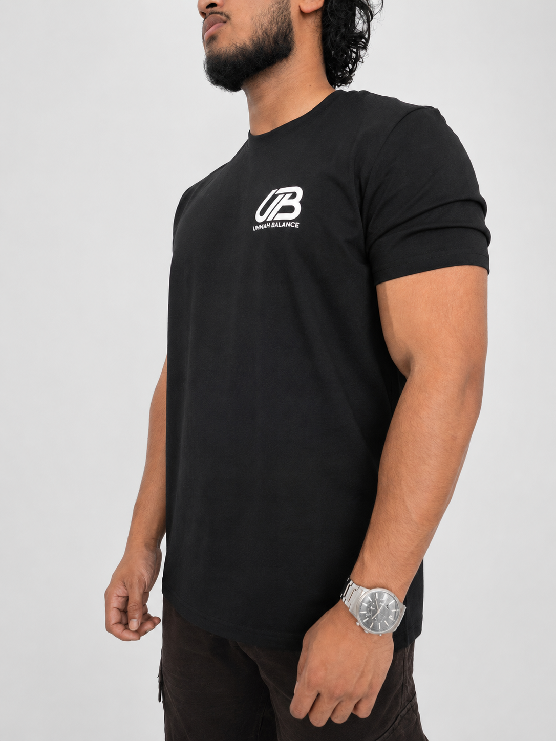 A man wearing a black short-sleeve t-shirt with a white 'UB' logo and 'Ummah Balance' text on the left chest, shown from the waist up against a light grey background.