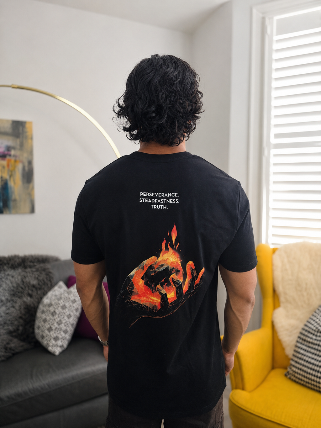 A man viewed from behind wearing a black t-shirt with a graphic of a hand holding a burning ember, with the words 'PERSEVERANCE. STEADFASTNESS. TRUTH'