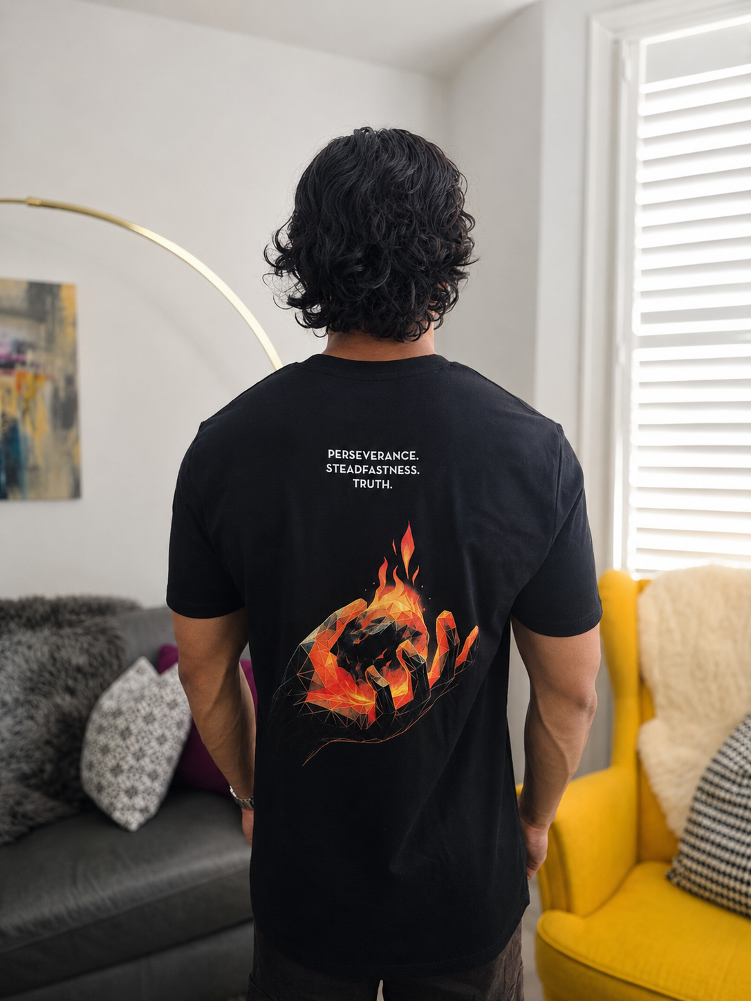 A man viewed from behind wearing a black t-shirt with a graphic of a hand holding a burning ember, with the words 'PERSEVERANCE. STEADFASTNESS. TRUTH'