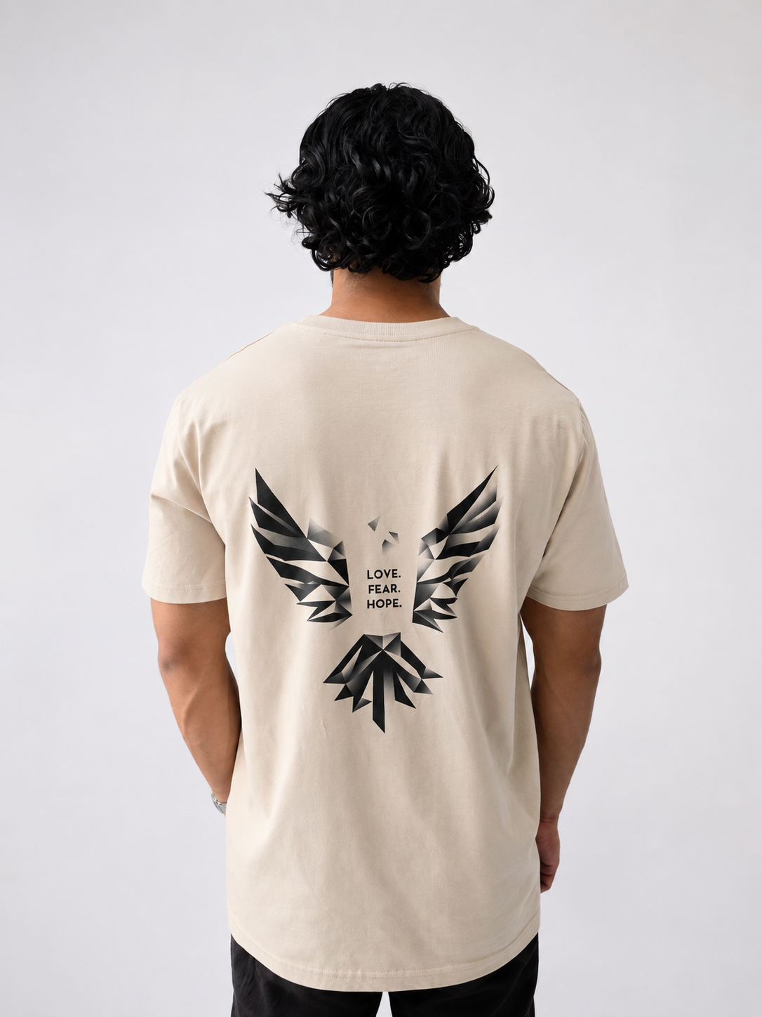 A man viewed from behind wearing a desert dust-coloured t-shirt with a large black geometric bird graphic and the words 'LOVE. FEAR. HOPE.'