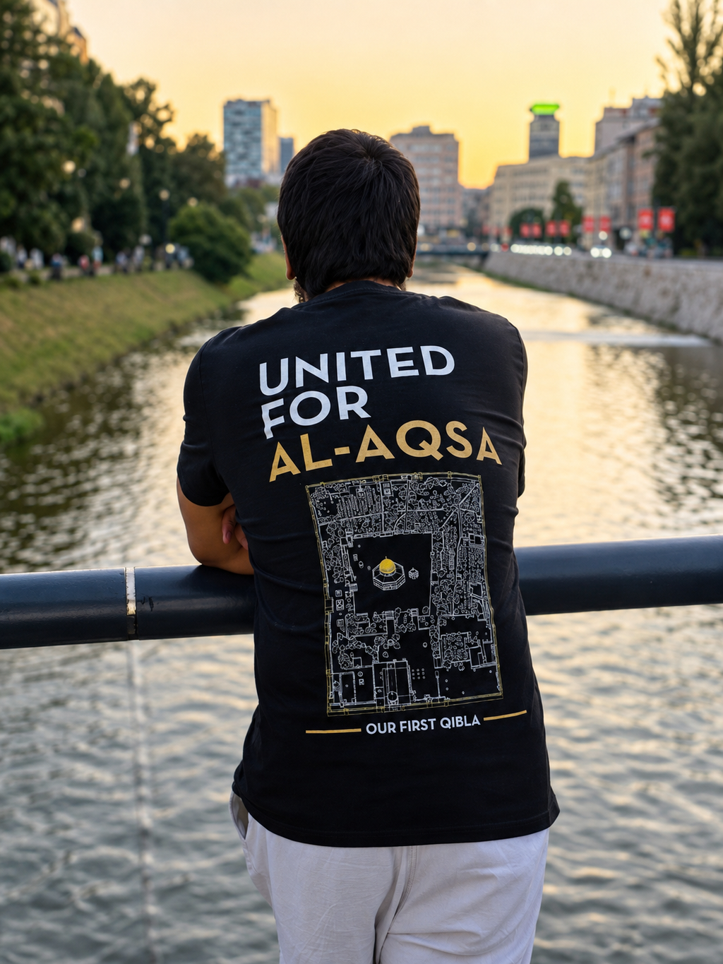 A man stands with his back to the camera, leaning on a bridge railing over a river at sunset, wearing a black t-shirt with a large graphic print on the back that reads 'UNITED FOR AL-AQSA' and 'OUR FIRST QIBLA'.
