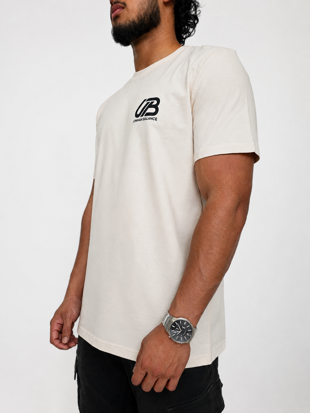 A man wearing a vintage white-coloured short-sleeve t-shirt with a black 'UB' and 'Ummah Balance' logo on the left chest, paired with black pants, against a plain white background.