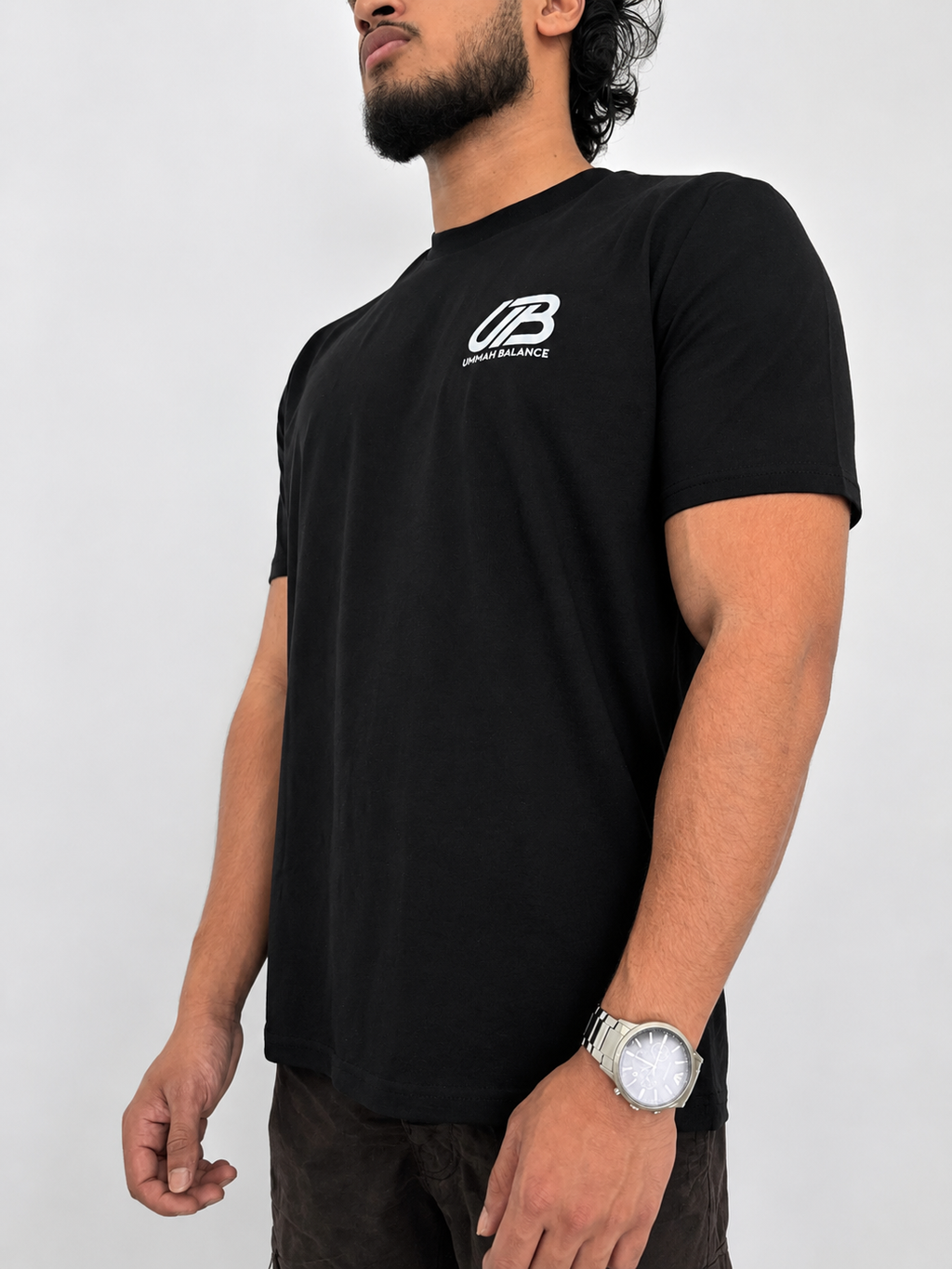 A man wearing a black short-sleeve t-shirt with a white 'UB' logo and 'Ummah Balance' text on the left chest, paired with dark brown pants and a silver watch, against a light grey background.