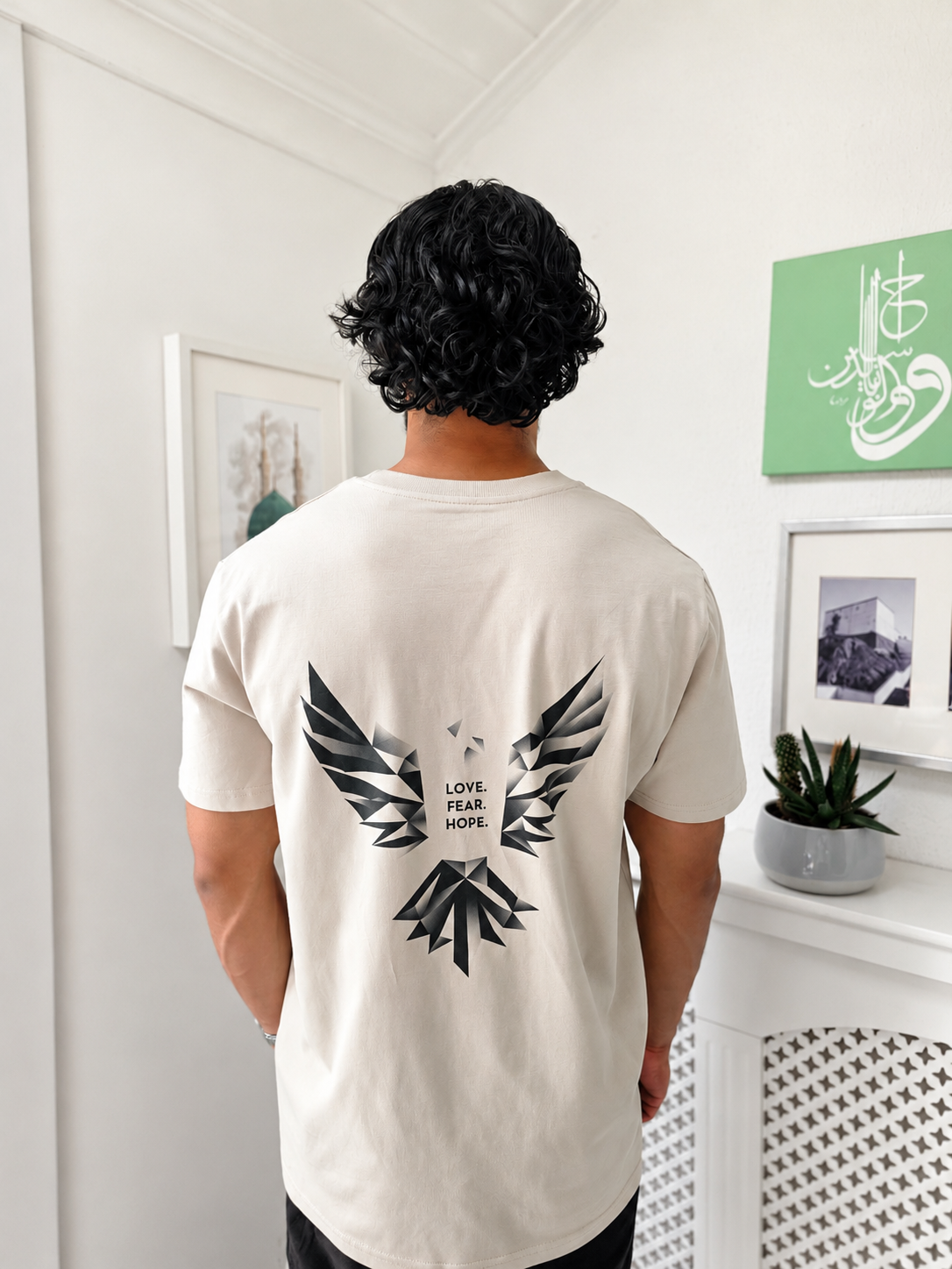 A man viewed from behind wearing a desert dust-coloured t-shirt with a large black graphic of a abstract bird and the words 'LOVE. FEAR. HOPE.' printed on the back, standing in a bright white room.