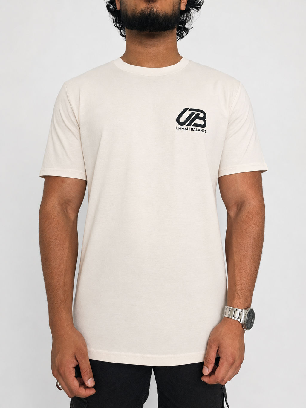 A man wearing a vintage white-coloured short-sleeve t-shirt with a black 'UB' and 'Ummah Balance' logo on the left chest, paired with black pants, against a light grey background.