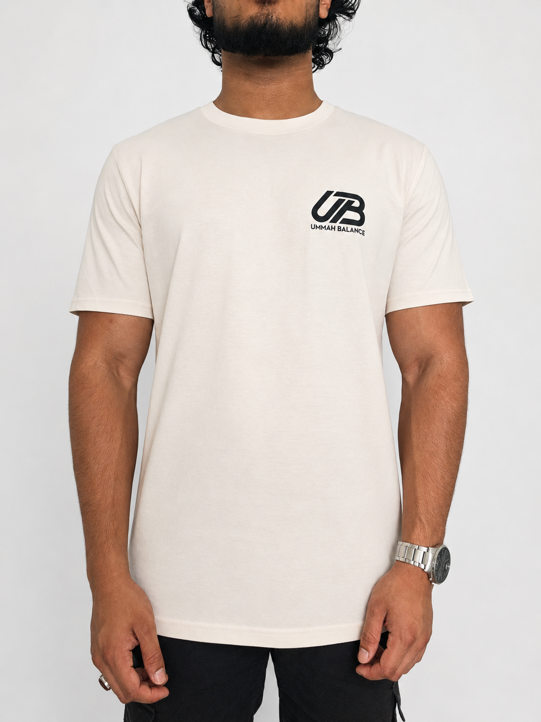 A man wearing a vintage white-coloured short-sleeve t-shirt with a black 'UB' and 'Ummah Balance' logo on the left chest, paired with black pants, against a light grey background.