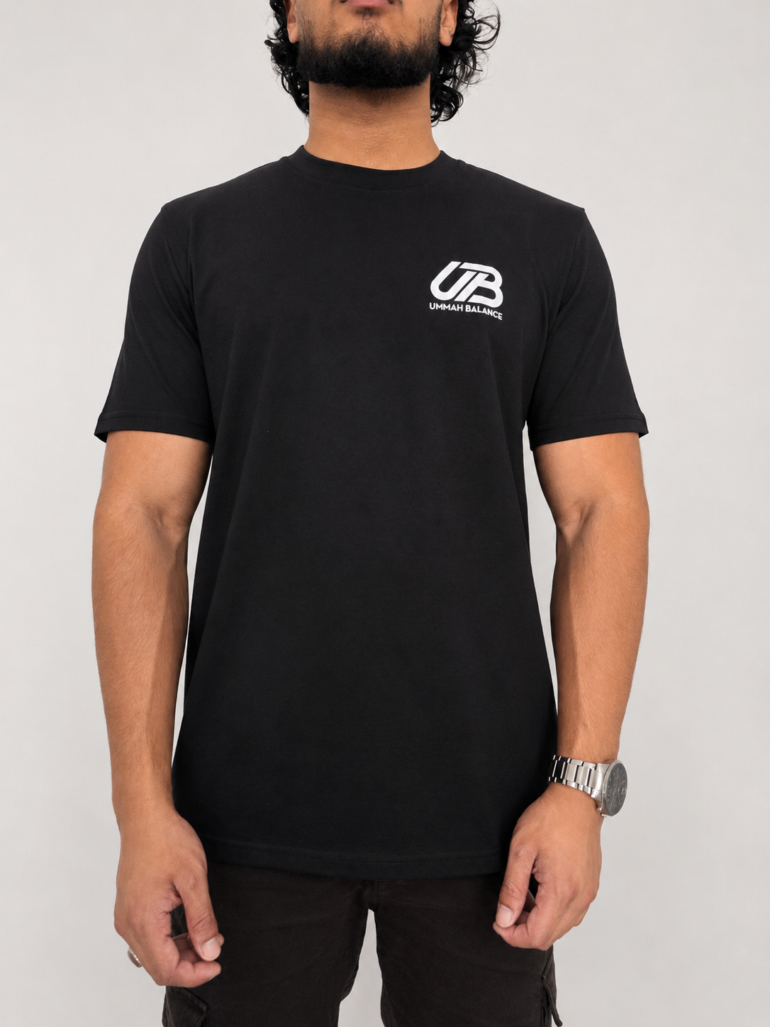 A man wearing a black short-sleeve t-shirt with a white 'UB' logo and 'Ummah Balance' text on the left chest, paired with black pants, against a light grey background.