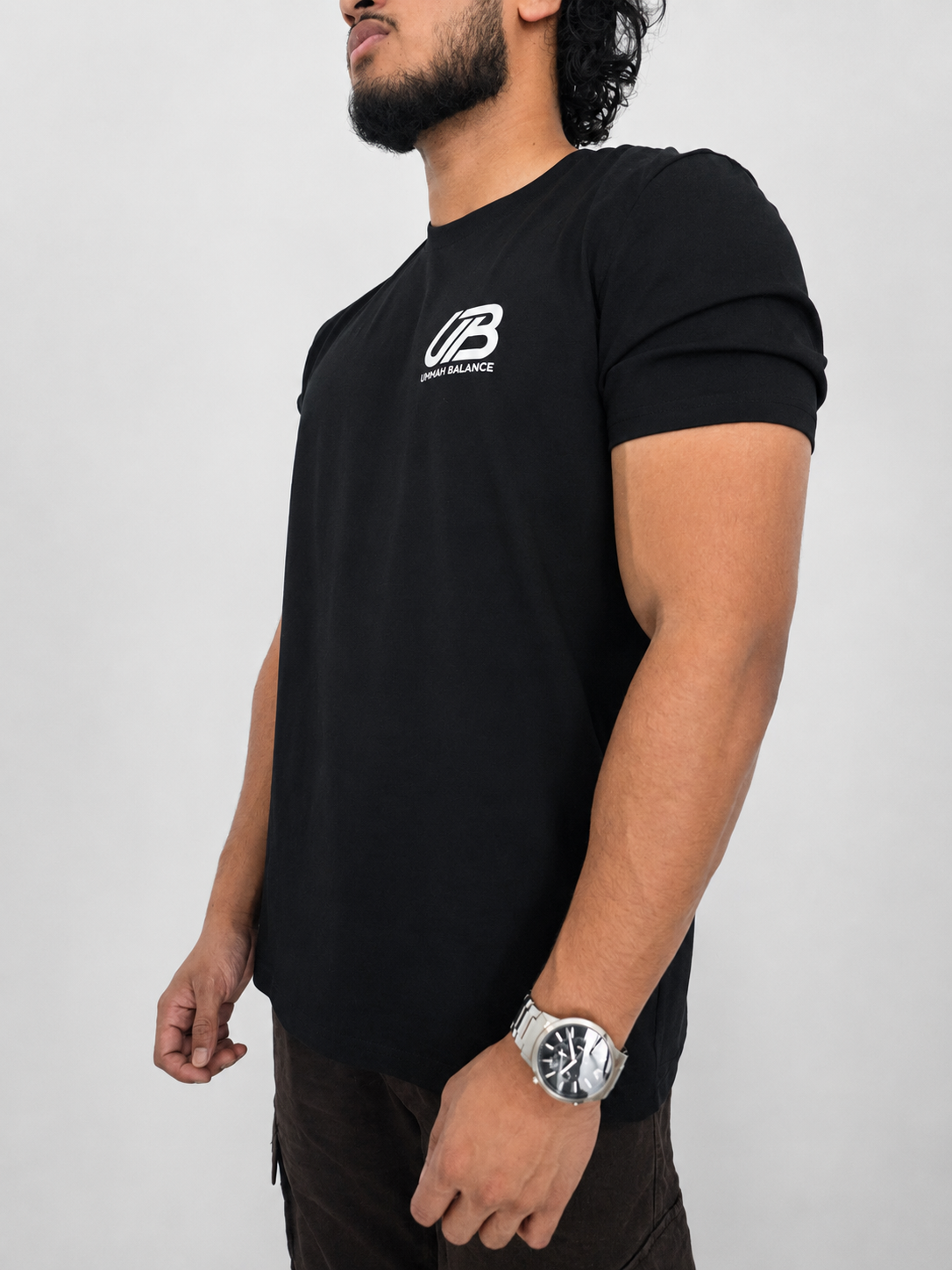 A man wearing a black short-sleeve t-shirt with a white 'UB' and 'Ummah Balance' logo on the left chest, shown from a side profile against a light grey background.