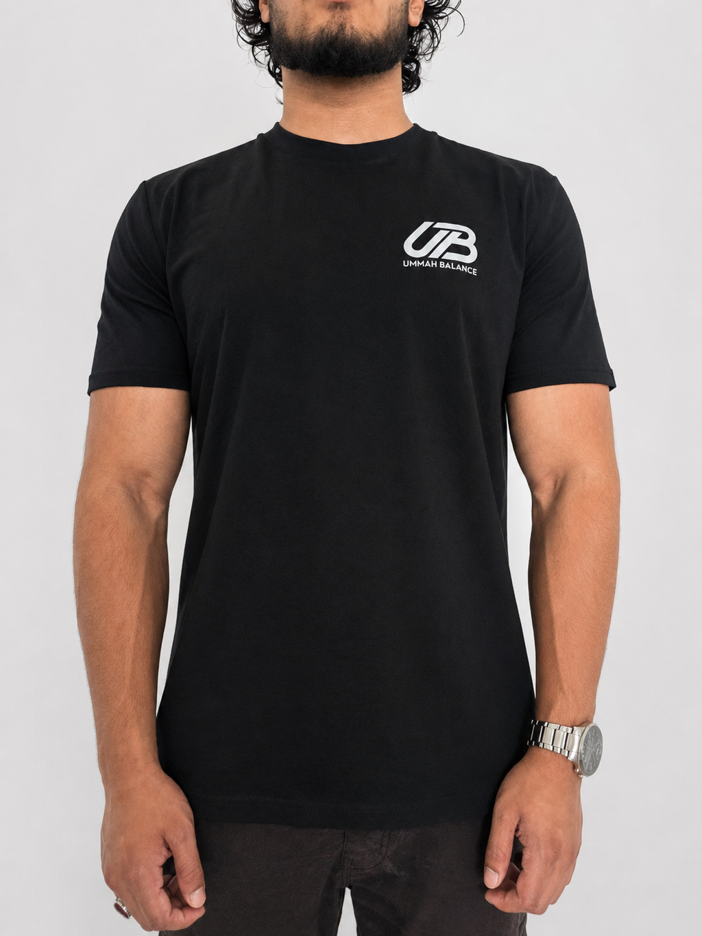 A man wearing a black short-sleeve t-shirt with a white Ummah Balance logo on the left chest, shown from the waist up against a light grey background.